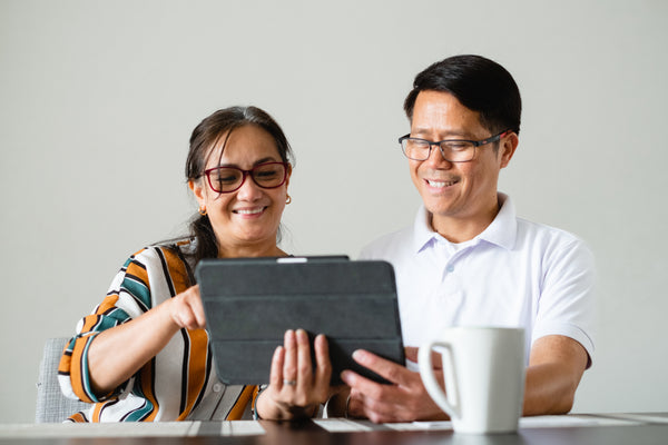 A couple smiling as they use a tablet together, enjoying their experience. The man has short dark hair and wears a white shirt, while the woman has long dark hair and wears a colourful top. The background is softly blurred, focusing on their happy interaction.