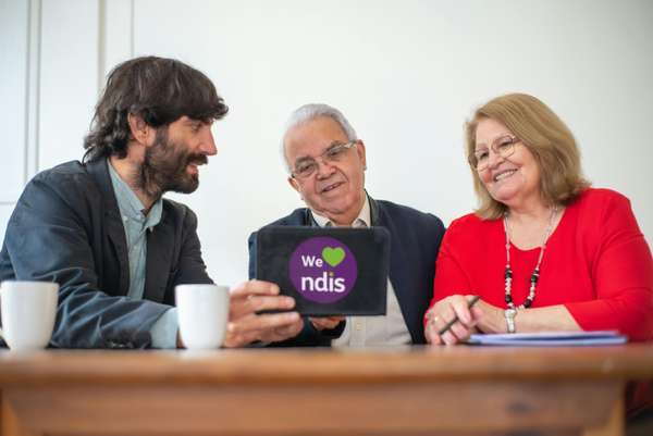 Three people at a table with cups and a notebook. The middle person holds a tablet showing “We ❤️ ndis” on a purple background. Left: dark jacket, blue shirt. Right: red top, beaded necklace. Bright and friendly setting.