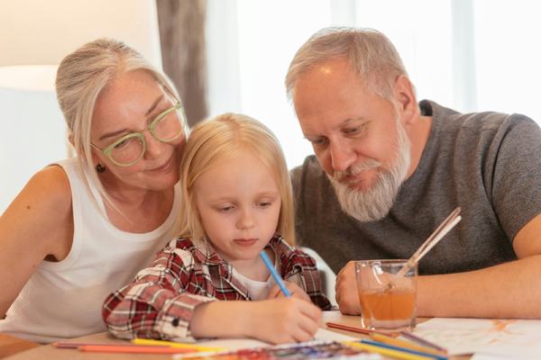 A child in a plaid shirt draws with a blue pencil, observed by two adults seated on either side. A glass of orange juice with a straw rests on the table. Colorful, engaging, and warm atmosphere.
