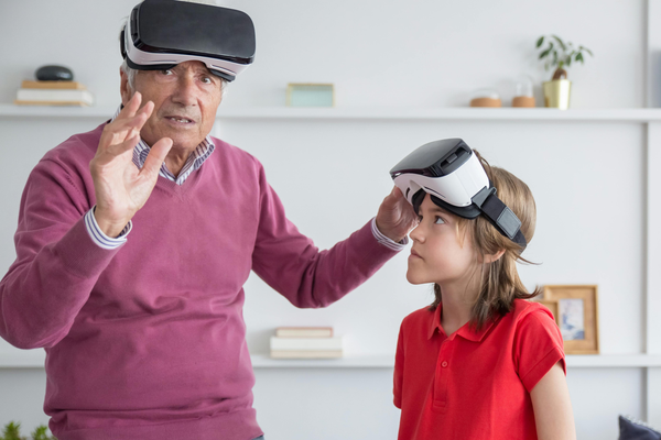 Grandfather and grandson wearing VR headsets. One in a pink sweater adjusts theirs, the other in a red shirt sits next to them. The background shows a bookshelf, a plant, and decorative items. Immersive atmosphere focusing on virtual reality.